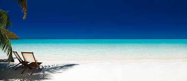 A panoramic shot of two teak folding chairs with white canvas seat backs and wooden seats on a tropical beach in St. John, US Virgin Islands. The chairs are next to each other and slightly angled on the far left of the image in the shadow of a palm tree which is just visible. The sand is white and fades into a turquoise sea with a clear deep blue sky.